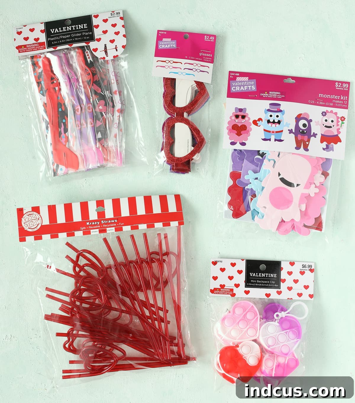 A flat lay photograph showcasing the five different non-food Valentine's Day card toys from Hobby Lobby: glider planes, glitter heart glasses, monster foam sticker kits, heart krazy straws, and heart fidget toy backpack clips.
