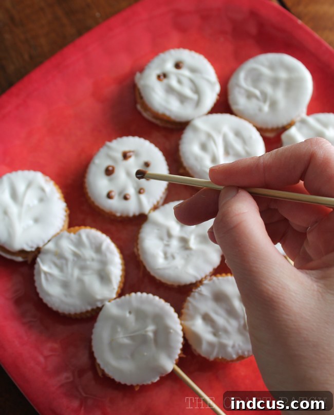 Close-up of a perfectly crafted Ritz cracker snowman snack on a stick, featuring intricate chocolate facial features and a distinctive orange frosting nose.