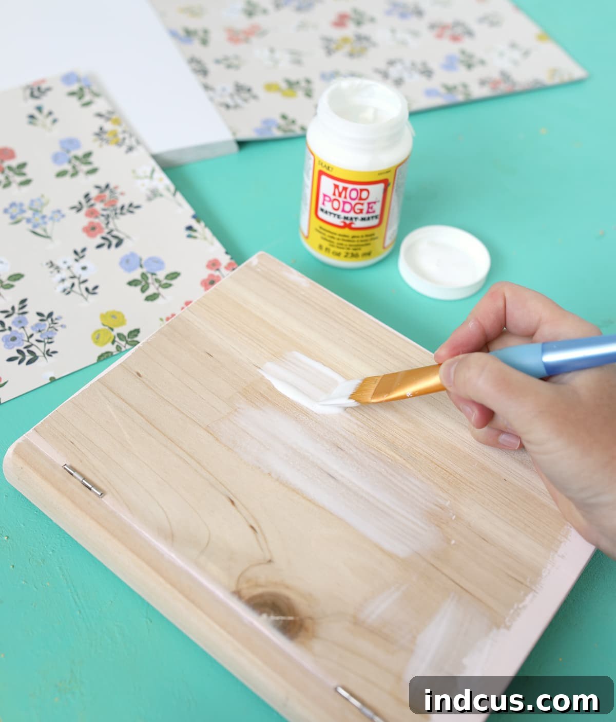 Crafting process showing Mod Podge being brushed onto a wooden box to adhere patterned paper, creating a decorative book box.
