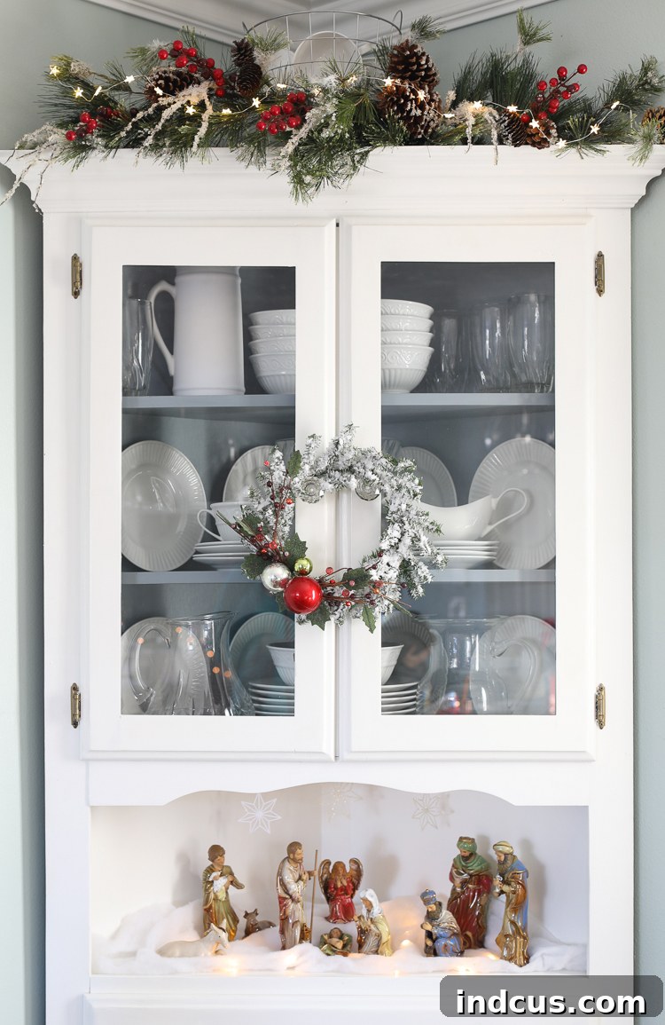 A beautifully decorated corner hutch for Christmas, featuring garland, fairy lights, and a nativity scene.