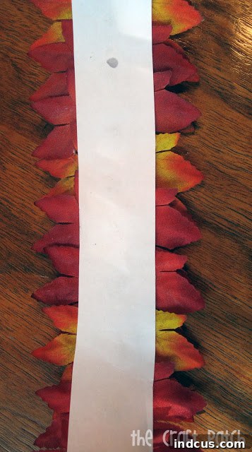 Cheap Chic Fall Decor 3 A step-by-step image showing how to hot glue faux fall leaves onto a strip of paper to create a DIY candle wrapper, perfect for autumn decorating.