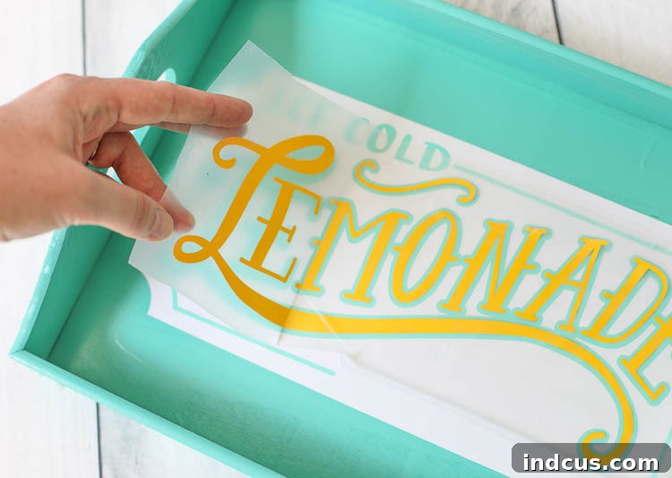 A close-up of a hand carefully peeling back transfer tape from a wooden tray, leaving behind a perfectly adhered white vinyl design layer.